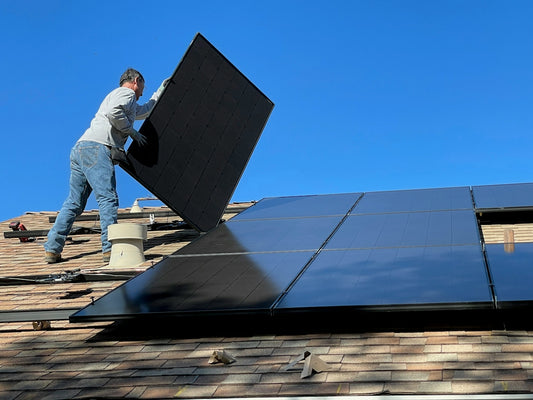A person installing a black solar panel on a shingled roof under blue sky